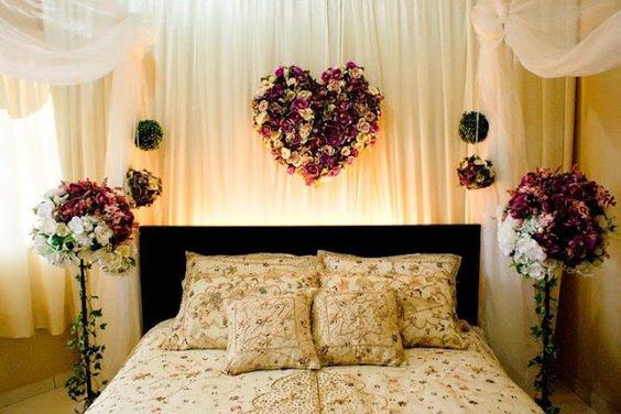 اتاق عروسی که در کنار و بالای تخت آن دسته گل های سفید و بنفش گذاشته شده است و تخت آن دارای روتختی طرح دار کرم رنگ می باشد